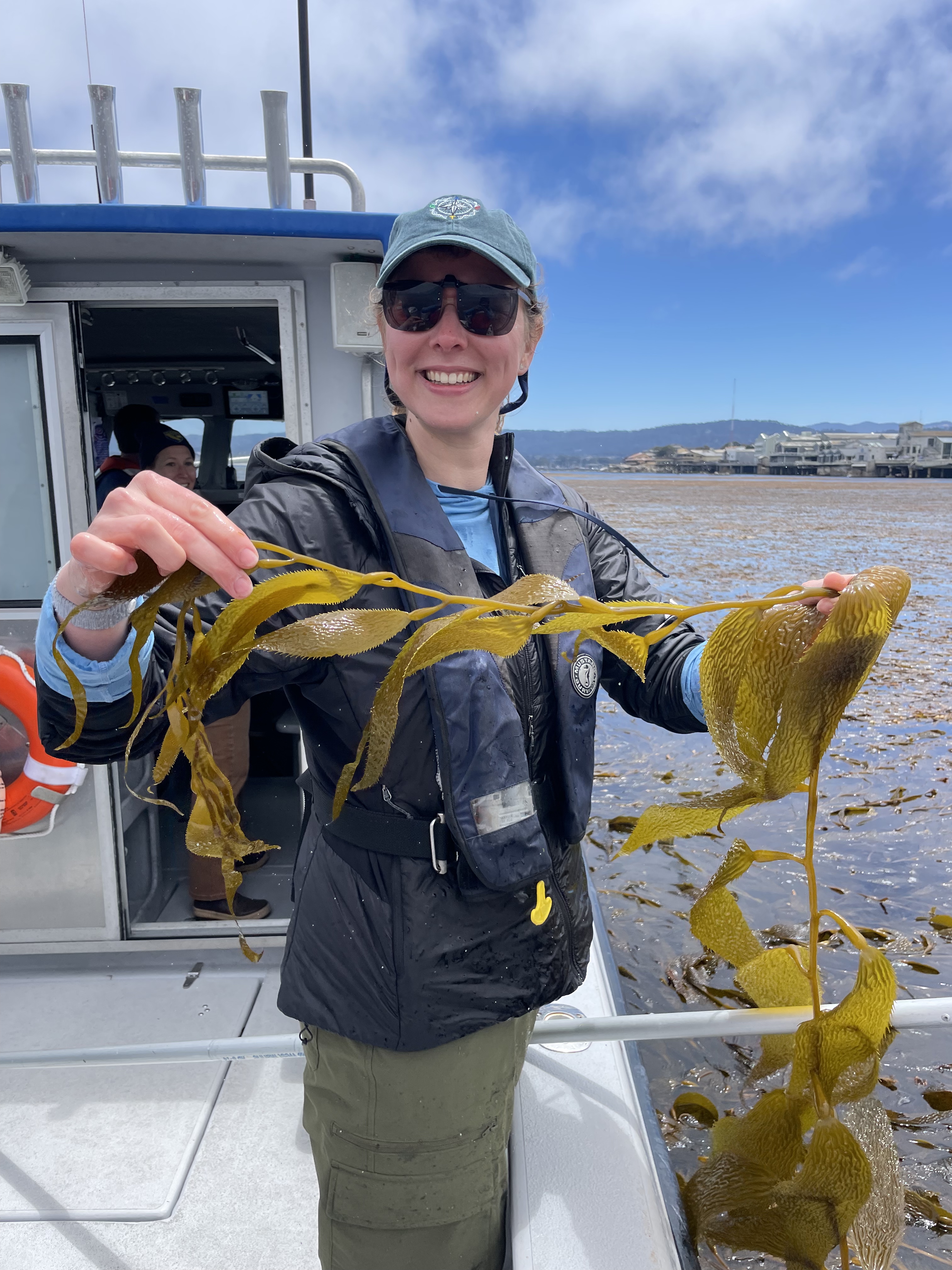 A woman smiling on a boat holding a strand of giant kelp up to the camera
